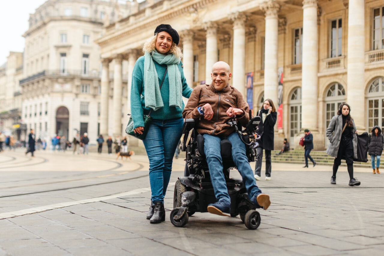 Mickael, souriant, atteint de l'infirmité motrice cérébrale assis sur son fauteuil roulant, à côté de son auxiliaire de vie dans les rues de Bordeaux