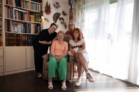 une bénéficiaire âgée est assise dans un fauteuil, entourée de trois accompagnantes dans un salon.