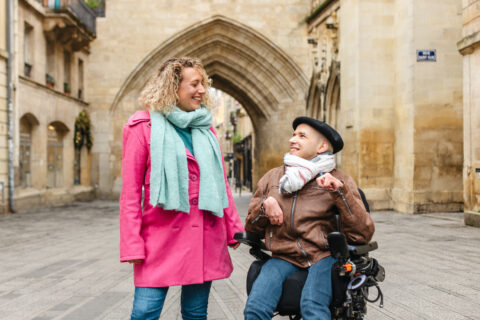 un beneficiaire en fauteuil roulant et son auxiliaire de vie se promènent et discutent en souriant dans une rue pavée bordée de bâtiments anciens