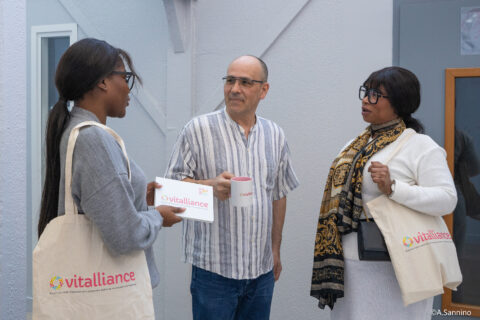 Trois adultes discutent à l’interieur ; une intervenante présente des documents Vitalliance à un couple, chacun portant un tote bag de la marque.